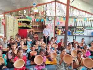 A group of children sit indoors, each holding a clay pot. Colorful decorations and posters adorn the background as an adult from the Shree Danev Foundation stands smiling among them.